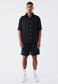 Zwarte T-shirt met korte mouwen en een gestructureerd patroon, voorzien van vijf witte knopen, gecombineerd met bijpassende shorts. Witte sneakers en sokken maken de outfit compleet.