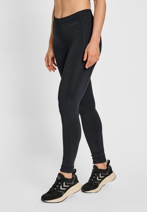 Kobieta ubrana w czarne dopasowane legginsy i czarno-białe sportowe buty, stojąca na jednolitym tle.