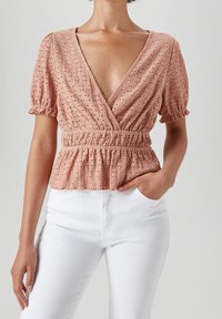 Blush-pink Peplum-Bluse mit V-Ausschnitt, kurzen Puffärmeln und strukturiertem Muster. Getragen mit weißen, hochtaillierten Jeans.