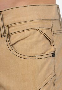 Pantalons kaki avec une surface texturée, des coutures noires marquées, et un design de poche classique. Comprend un détail de bouton visible.
