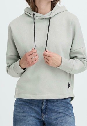 Person trägt hellgrauen Hoodie mit schwarzen Kordeln und blaue Jeans, hält die Hoodie-Kordeln mit beiden Händen nahe der Brust.