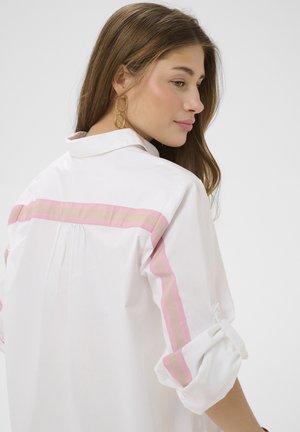 Ung kvinde med langt brunt hår ser til siden, iført dobbelte gyldne hoop-øreringe og en hvid skjorte med lyserøde og beige stribede detaljer.