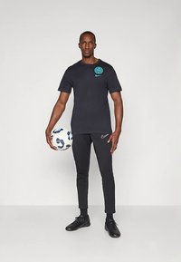 Mustasportlik mees hoiab valget ja sinist jalgpalli, seistes ühetoonilise heleda halli tausta ees.