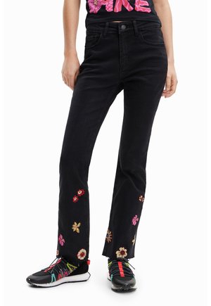 Person iført sorte jeans med flerfarvet blomsterbroderi nær kanten, kombineret med sorte og farverige sneakers.