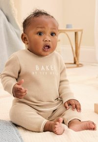 Ensemble pour bébé en tricot beige avec des manches longues et des poignets, arborant l'inscription "BAKER BY TED BAKER". Posé sur une surface légère et texturée.