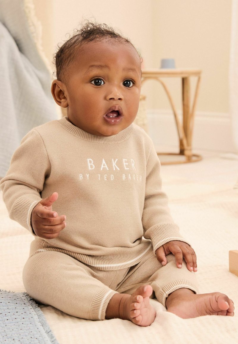 Ensemble pour bébé en tricot beige avec des manches longues et des poignets, arborant l'inscription "BAKER BY TED BAKER". Posé sur une surface légère et texturée.