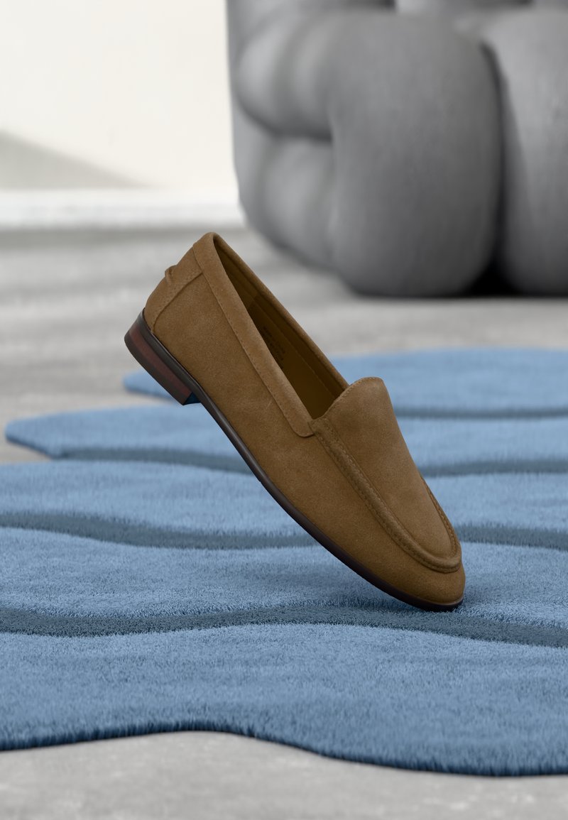 Mocassin en daim marron posé sur un tapis bleu texturé avec une grande sculpture grise floue en arrière-plan.