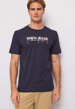 Uomo con maglietta blu navy con testo colorato "GAUDI JEANS" e pantaloni beige, in piedi davanti a uno sfondo chiaro uniforme.