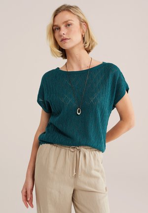 Plavolasa ženska, oblečena v turkizno teksturirano majico s kratkimi rokavi, bež hlače z vrvico in obesek na vratu, stoji ob nevtralnem ozadju.