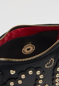 Černá kožená kabelka se červeným podšívkováním, zlatými prvky, srdíčkovými cvoky a logem ve tvaru srdce na zipu.