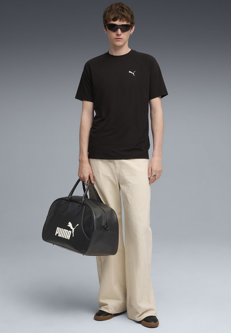Sort Puma T-shirt med logo, cremede vidde bukser, holder en sort læder duffeltaske med et hvidt logo og bærer solbriller.