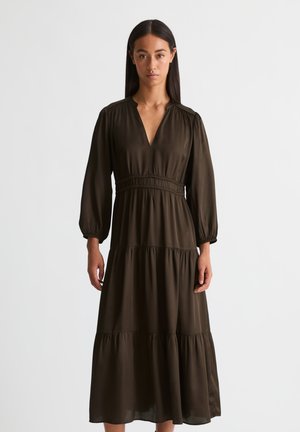 Frau mit langen dunklen Haaren, die ein langärmliges, dunkelbraunes, gestuftes Maxikleid mit V-Ausschnitt trägt und vor einem schlichten weißen Hintergrund steht.