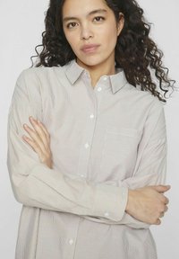 Camisa de botones de rayas de color claro con cuello y mangas largas, que presenta un bolsillo en el lado izquierdo del pecho y botones blancos.