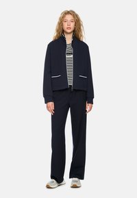 Navy athletic jacket with white piping, paired with a striped shirt and navy pants. Footwear features a mix of white and gray tones.