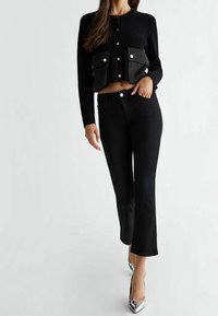 Kvinde iført sort kort cardigan med store forlommer, sorte højtaljede jeans og sølvfarvede spidse hæle, stående mod en hvid baggrund.