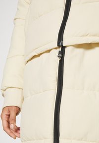 Primer plano de una persona que lleva una chaqueta acolchada color crema con una cremallera frontal negra y mangas de puño.