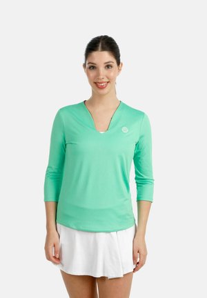 Mujer sonriendo, vestida con un top de manga larga verde menta con un pequeño logo y una falda plisada blanca, de pie frente a un fondo liso.