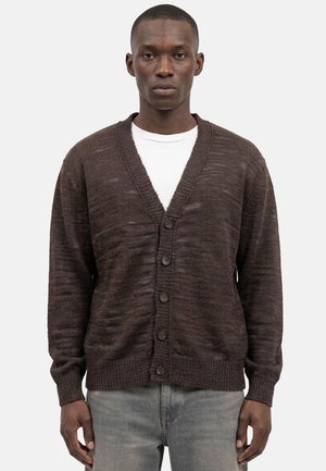 Uomo che indossa un cardigan marrone scuro abbottonato sopra una camicia bianca e jeans grigi, in piedi davanti a uno sfondo chiaro e neutro.