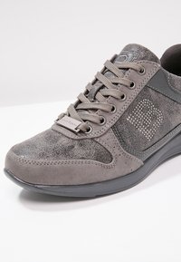 Chaussure de sport grise en matériau synthétique texturé, avec une coque d'orteil lisse, des lacets et des strass décoratifs sur le côté.
