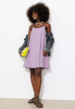 Femme souriante aux cheveux afro portant une robe lavande, une veste en jean tombant des épaules, des colliers en coquillages, tenant une pochette jaune et des sandales noires.