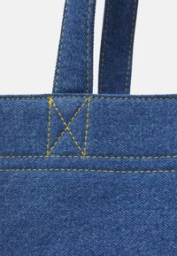 Sangle de sac en denim bleu attachée avec une couture renforcée jaune sur fond blanc, montrant la texture du tissu et les détails des coutures.