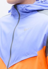 Ανοιχτό μπλε και πορτοκαλί μπουφάν με φερμουάρ και κουκούλα, με το λογότυπο της Nike στο στήθος, φορεμένο από άτομο που φαίνεται από το λαιμό έως το μέσο του κορμού.