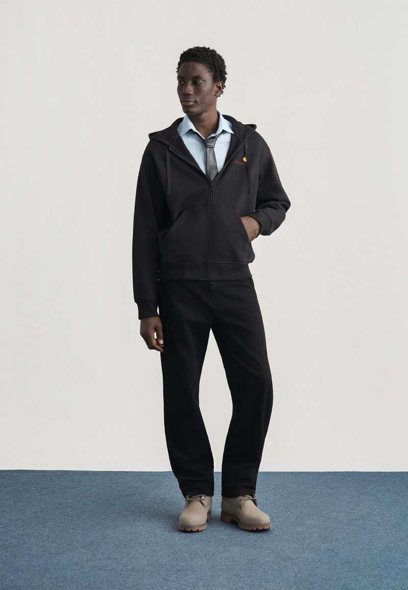 Junger Mann steht auf blauem Teppich, trägt schwarzen Hoodie, hellblaues Hemd mit grauer Krawatte, schwarze Hose und beige Stiefel, blickt seitlich.