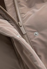 Bézs színű kabát belső része, amelyen jól látható cipzár és patentgomb található, texturált felülettel és finom négyzetmintázattal a szöveten.