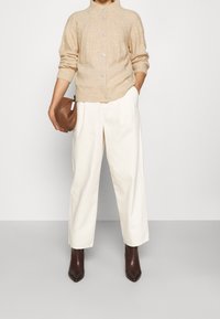 Cardigan en maille côtelée beige clair avec un col montant et des boutons argentés, associé à un pantalon large crème et des bottes pointues marron foncé.
