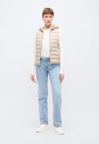 Μπεζ αμάνικο μπουφάν πάπλωμα με κουκούλα, με ραμμένο σχέδιο. Φορεμένο πάνω από ένα λευκό μακρυμάνικο μπλουζάκι και συνδυασμένο με ανοιχτόχρωμο τζιν και λευκά αθλητικά παπούτσια.