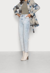 Mujer que lleva jeans de tiro alto de lavado claro, botas de tacón beige hasta el tobillo, top verde oliva y un abrigo de color gris-azul con patrón, con las manos en las caderas.