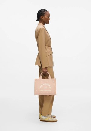 Mujer con trenzas en un traje beige sosteniendo un bolso tote rosa de Karl Lagerfeld, de perfil sobre un fondo blanco.
