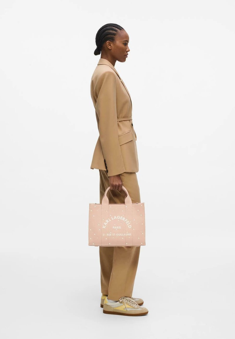 Femme aux cheveux tressés en costume beige tenant un sac cabas rose Karl Lagerfeld, debout de profil sur fond blanc.