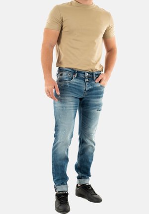Camiseta ajustada beige combinada con jeans azul ligeramente deslavado que presentan detalles desgastados, complementada con zapatillas negras de perfil bajo.
