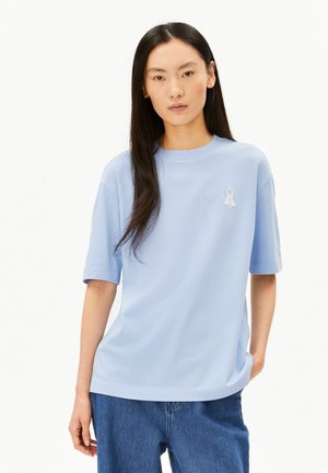 Young woman with long dark hair wearing a light blue short-sleeve shirt and blue jeans against a plain white background.