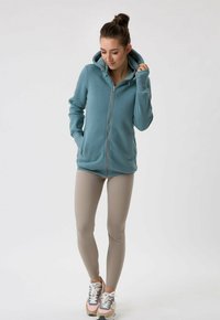 Felpa azzurra con zip, dotata di un cappuccio ampio, tasche frontali e orlo a coste. Abbinata a leggings beige e scarpe da ginnastica multicolori.