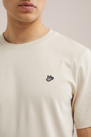 Detailná fotografia osoby, ktorá má na sebe béžové tričko s malým čierno-bielym logom vtáka vyšitým na ľavej hrudi.
