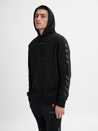 Jaunas vyras dėvi juodą gobtuvu megztinį ir derančias juodas kelnes su strėlių raštais ant rankovių ir kelnių kojų, stovintis prieš baltą foną.