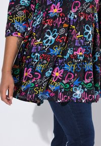 Haut en coton coloré avec une base noire et des motifs graphiques multicolores, y compris des fleurs, des flèches et du texte. Coupe décontractée à volants.