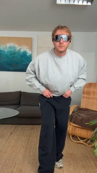 Grå oversized hettegenser, svarte joggebukser og reflekterende solbriller. Myk tekstur og avslappet passform. Lys bakgrunn med abstrakt kunst.