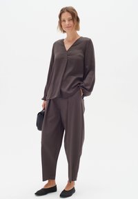Dark brown, loose-fitting blouse with a V-neck and long sleeves, paired with matching tapered pants and black ballet flats. Holding a small black bag.