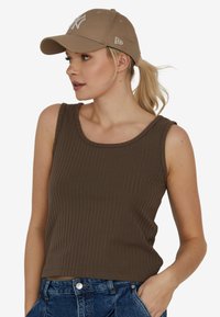 Kobieta z blond kucykiem, nosząca brązowy prążkowany top bez rękawów, beżową czapkę bejsbolową i niebieskie dżinsy, z ręką w kieszeni, patrząca w lewo.