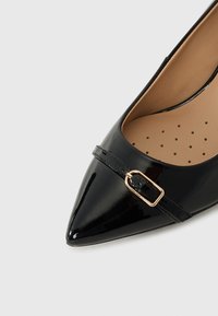 Zapato de tacón alto de charol negro con punta afilada, que presenta una correa delgada con una hebilla dorada y un forro interior perforado.