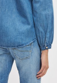 Lyseblå denimskjorte med avrundet kant, knappede mansjetter og glatt tekstur. Kombinert med matchende lyseblå jeans med lomme.