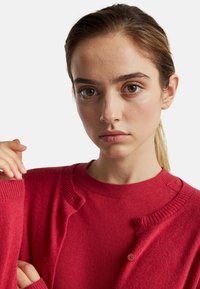 Červený kardigan vyrobený z měkkého materiálu, s knoflíkovým zapínáním a žebrovanými manžetami. Nositelný přes odpovídající červený svetr. Mírná textura viditelná.