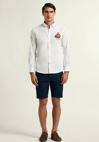 Camisa blanca de manga larga con cierre de botones, que presenta un logo bordado. Combinada con pantalones cortos azules y zapatos marrones. Diseño simple, sin patrones.