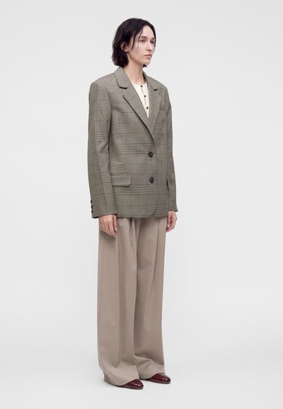 Femme debout portant un blazer à carreaux marron oversize, une chemise blanche boutonnée, un pantalon beige large et des chaussures rouge foncé sur un fond uni.