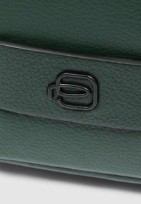 Grüne Ledertasche mit strukturierter Oberfläche und einem auffälligen schwarzen Logo-Akzent. Das Design umfasst eine schlanke, rechteckige Form.