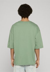 Bledě zelené oversized tričko s krátkými rukávy, kulatým výstřihem a hladkou texturou. Zadní pohled ukazuje uvolněný střih a boční rozparky.