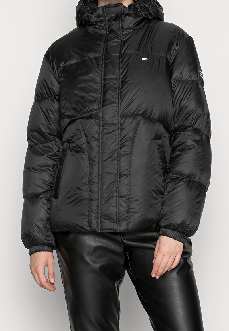 Zwarte pufferjack met een capuchon, voorzien van een voorrits, elastische manchetten en zijzakken. Gladde textuur en geborduurd ontwerp.